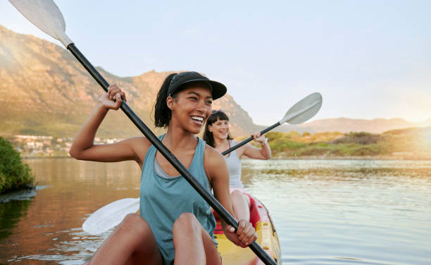 夏休みに一緒に湖でカヤックをしている笑顔の友人2人。笑顔で幸せな遊び心のある女性は、自然の中で水遊びと外で結びついています。週末のレクリエーション中にカヤックで楽しむ - recreational pursuit carefree nature vacations ストックフォトと画像