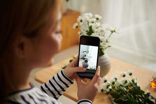 Closeup image of young woman photographing daisy flowers in vase on smartphone