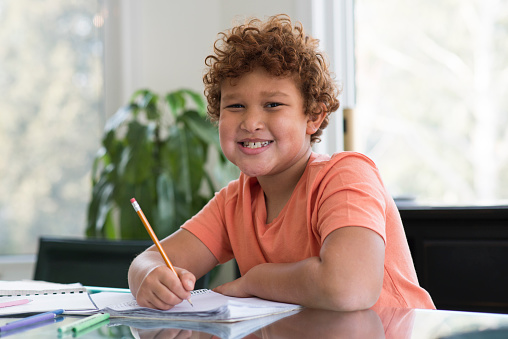 Young elementary age boy is smiling as he is looking at the camera and sitting at the kitchen table with a pencil in his hand doing school work from home.