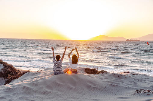 Two young adult women sitting on the beach with their arms raised and watching an amazing sunset.