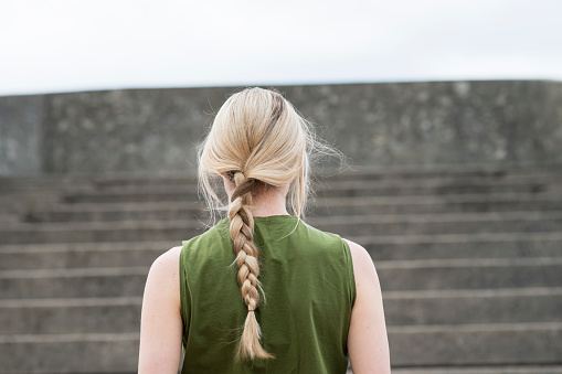 Anonymous young woman with her long blonde hair in a braid is facing away from the camera looking up a tall set of stairs in preparation for running up them. She is training her cardio and endurance while doing a workout outdoors. She is wearing a green sleeveless top.