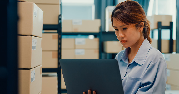 Asian businesswoman stand front of shelf use barcode machine scan customer data in paper box register online code order detail in laptop for express delivery at warehouse. Startup business concept.