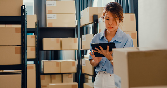 Asian businesswoman standing front of shelf use digital tablet check stock on shelf and key data in online information detail in before express delivery at warehouse. Startup small business concept.