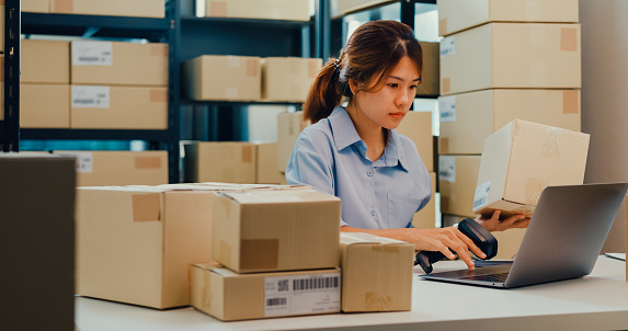 Asian businesswoman sit front of shelf use barcode scan customer data in paper box on desk register online code order detail in laptop for express delivery at warehouse. Startup business concept.
