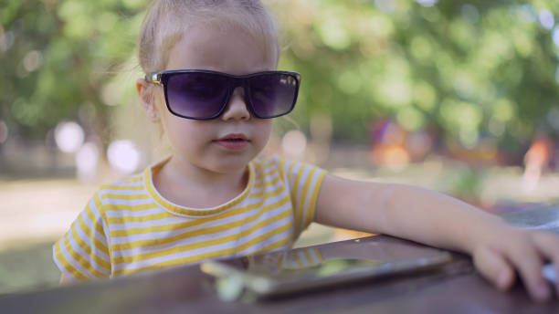 Little girl in mom's sunglasses is learning foreign language by repeating words from mobile phone. Close-up portrait of child girl sitting in city park and learning foreign language using cellphone la niña con gafas de sol de mamá está aprendiendo un idioma extranjero repitiendo palabras desde el teléfono móvil. retrato en primer plano de una niña sentada en el parque de la ciudad y aprendiendo un idioma extranjero usando el teléfono celular