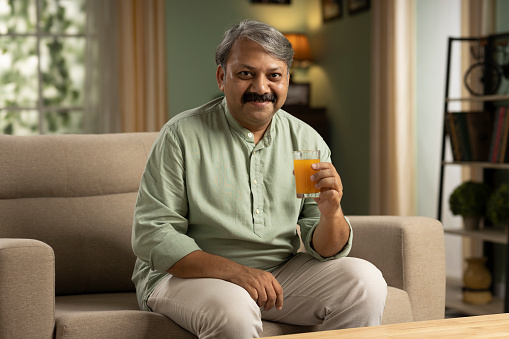 Happy senior man sitting on sofa at home and drinking orange juice