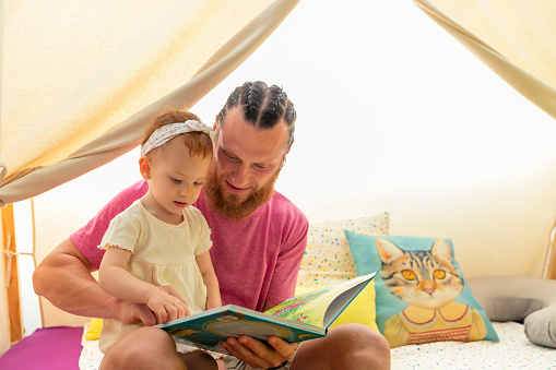 father with red-haired beard reading a story with his young daughter in the playroom