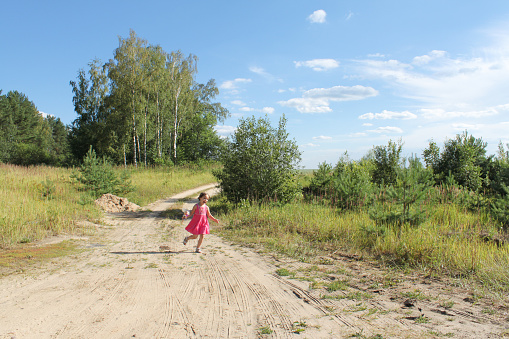 Funny toddler girl in pink dress with a doll runs along sandy path among fields and forest. Copy space.