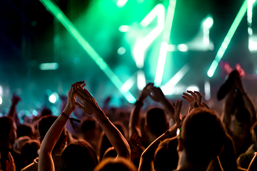 Rear view of a large group of people in front of a music festival stage. Crowd is excited and dancing, raising hands, clapping, punching the air, filming with mobile phones, etc...