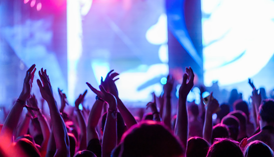 Rear view of a large group of people in front of a music festival stage. Crowd is excited and dancing, raising hands, clapping, punching the air, filming with mobile phones, etc...