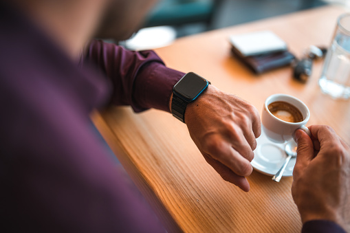 Caucasian man using a smart phone and a smart watch while drinking coffee in a cafe. He is checking his emails and calendar.