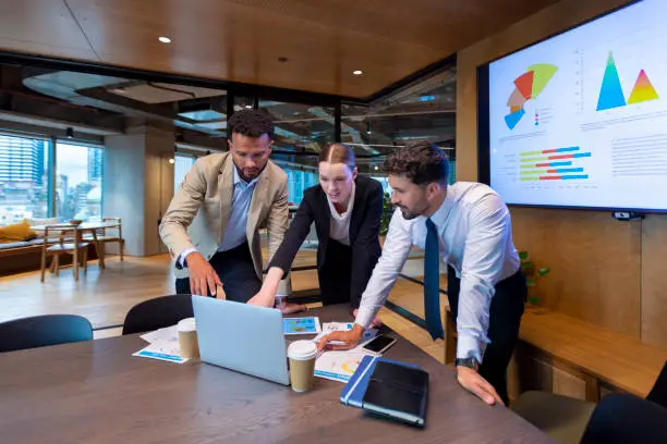 Business people working on a laptop computer in a modern office. There is paperwork on the table with charts and graphs on it. There is a screen behind them with financial charts and graphs on it.