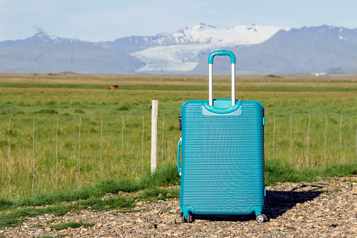 Single blue baggage suitcase standing against green pasture and mountains with glacier on sunny day and blue sky. Lot of copy space on side. Iceland nature.