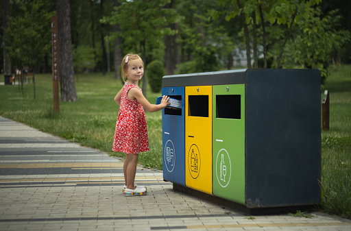 Cute little girl throw paper garbage into blue container in a park. Garbage sorting set. Bins with symbols for glass, paper and plastic trash. Care of environment by waste sorting
