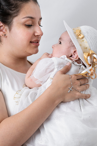 beautiful young latin mother, holding her baby with a silver chain entangled in her hand and with a cross pendant. baby baptism in catholic church. concept of religion
