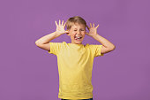 Young blond Caucasian kid preteen boy making faces staying against purple background. Studio shot.