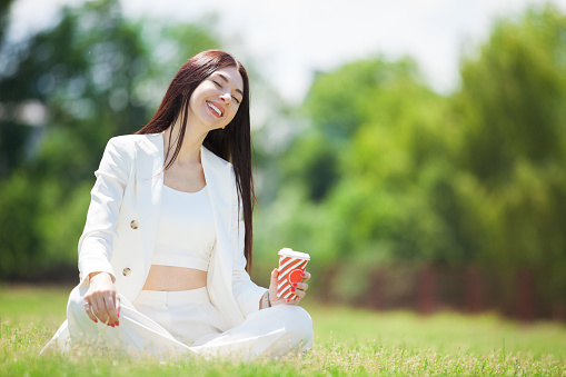 Beautiful business woman relax in park outdoors. Young cute lady in white suit sitting on the green grass and drink coffee. And let the whole world wait.