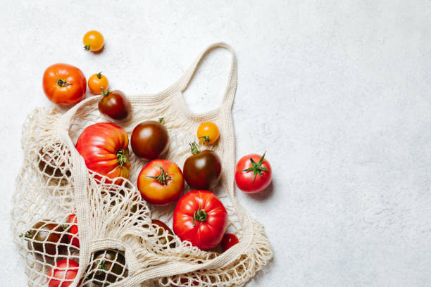Fresh colorful heirloom veriety tomatoes in eco friendly mesh shopping bag on white concrete background. Zero waste stock photo