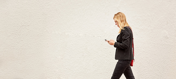 Side view of a businesswoman using her mobile phone while walking in front of a gray wall