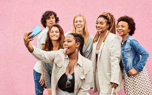 Group of multi-ethnic friends standing together and taking funny selfies against pink background