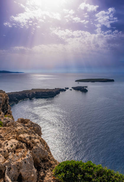 seascape from rocky cliffs hours before sunset, sunbeams filtering through small clouds, vertical, Fornells, Menorca, Balearic Islands, Spain