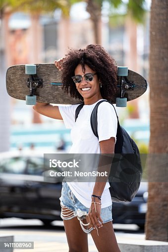 istock Young afro woman smiling and posing with skateboard. Sport female model in urban style. Street style outfit. Hipster girl skateboarding. 1411108960