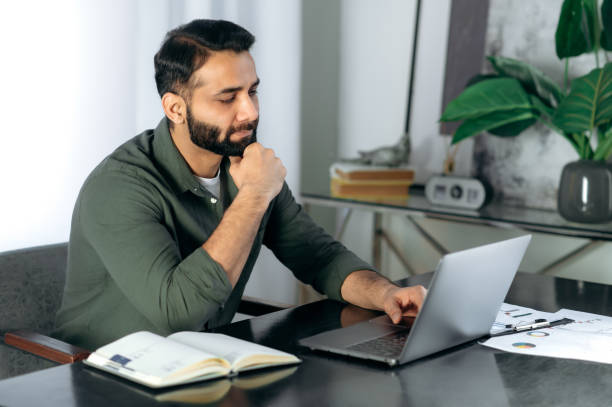 istock Conceived serious bearded indian or arabian guy in casual clothes, office worker or freelancer, sits at the desktop in the modern office, looks into the laptop intently, thinks about the project 1411109258