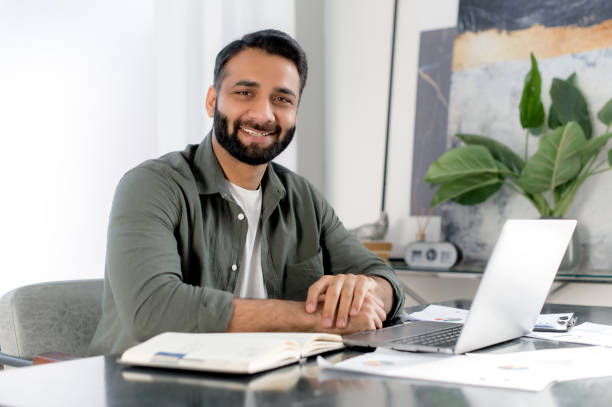 istock Positive confident indian or arabian man using laptop in modern office. Portrait successful businessman, freelancer, manager, sits at workplace in front of laptop, looking at camera, smiling friendly 1411109273