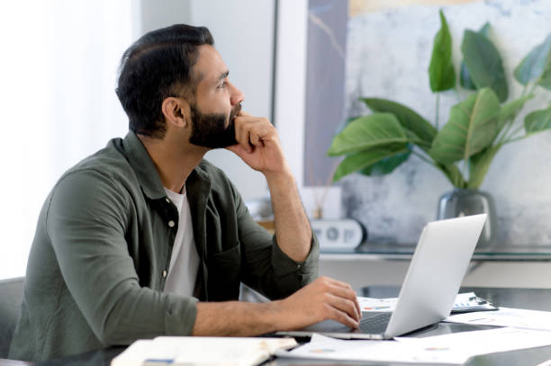 istock Pensive successful mixed race man, office worker, sitting in a modern office, distracted from work in a laptop, looking away thoughtfully, thinking about a new project, dreaming about vacation 1411109282