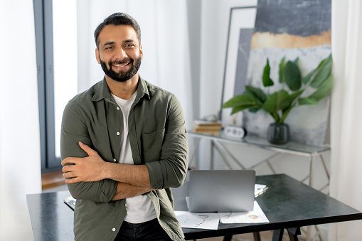 Portrait of a successful confident Indian man, creative manager, IT specialist, designer, wearing casual wear, standing near desk in the office with arms crossed, looks at the camera, friendly smiling