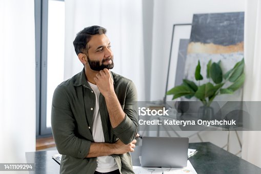 istock Portrait of a successful stylish Indian or Arabian businessman, manager or IT specialist, wearing casual stylish clothes, stand near his desk in the office with arms crossed, looking away and smiling 1411109294