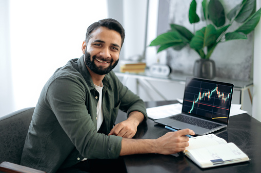 Portrait of a positive successful Indian or Arabian trader, stock market broker, investor, sitting at a desk in the office, looking at the camera, smiling friendly. Crypto charts on laptop screen