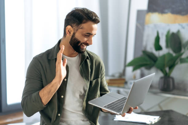 istock Successful arabian or indian man, holds and using laptop while stand in modern office, talking with coworker or client via video conference by laptop, conducts virtual meeting, smiling friendly 1411109300