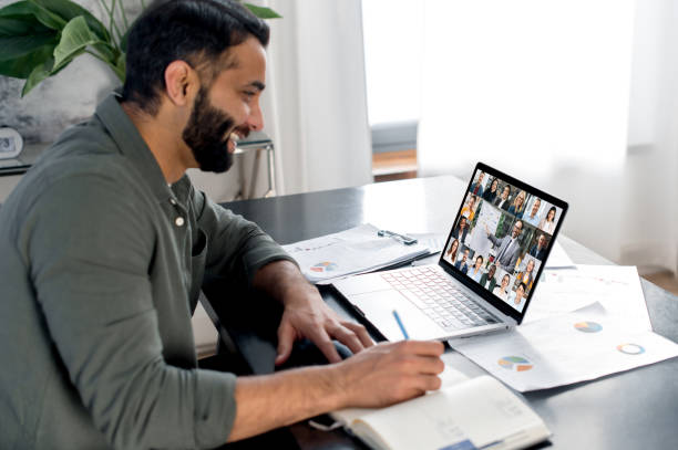 istock Brainstorming, briefing, business webinar. Smart man sit at a table, uses laptop, listens to a financial lecture, increases financial knowledge, makes notes in a notebook, multiracial people on screen 1411110337