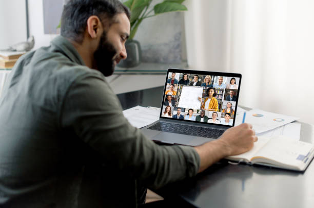 Online training, webinar. Over shoulder view of a successful smart guy listening to an online lecture, taking notes in a notebook, on a laptop screen, a teacher and a group of multiracial people stock photo