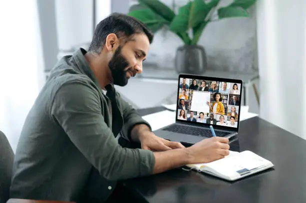 Photo of Side view of a successful smart guy listening to an online lecture, taking notes in a notebook, on a laptop screen, a teacher and a group of multiracial people. Online training, webinar