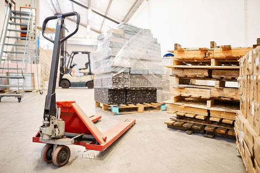 Pallet jack and boxes on pallets standing on the floor of a large manufacturing and shipping warehouse