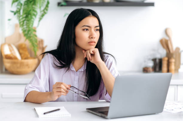 istock Thoughtful Chinese girl, freelancer, student, content manager, working from home, sit at a desk with a laptop in the kitchen, thoughtfully looking straight, thinks over a project plan, making decision 1411113982