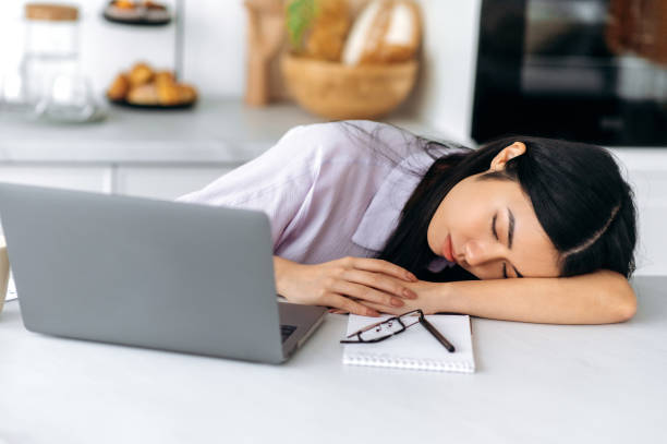 giovane donna di etnia cinese assonnata, si addormenta mentre è seduta sul posto di lavoro vicino al laptop a casa in cucina, stanca di lavori noiosi, oberata di lavoro, che sperimenta la privazione cronica del sonno, ha bisogno di riposo - yawning women drink coffee cup foto e immagini stock