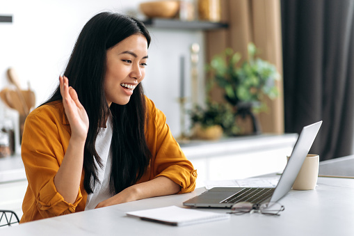 Side view of an Asian positive sociable girl, in casual clothes, working from home sitting in the kitchen, communicates via online video conference with employees, greets, smile, discusses work issues