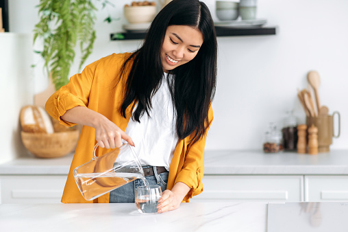Positive Asian girl, stands in the kitchen, pouring fresh clean water from a jug into a glass, care her health, replenishes energy and strength in the body, drink the daily amount of water, smiling