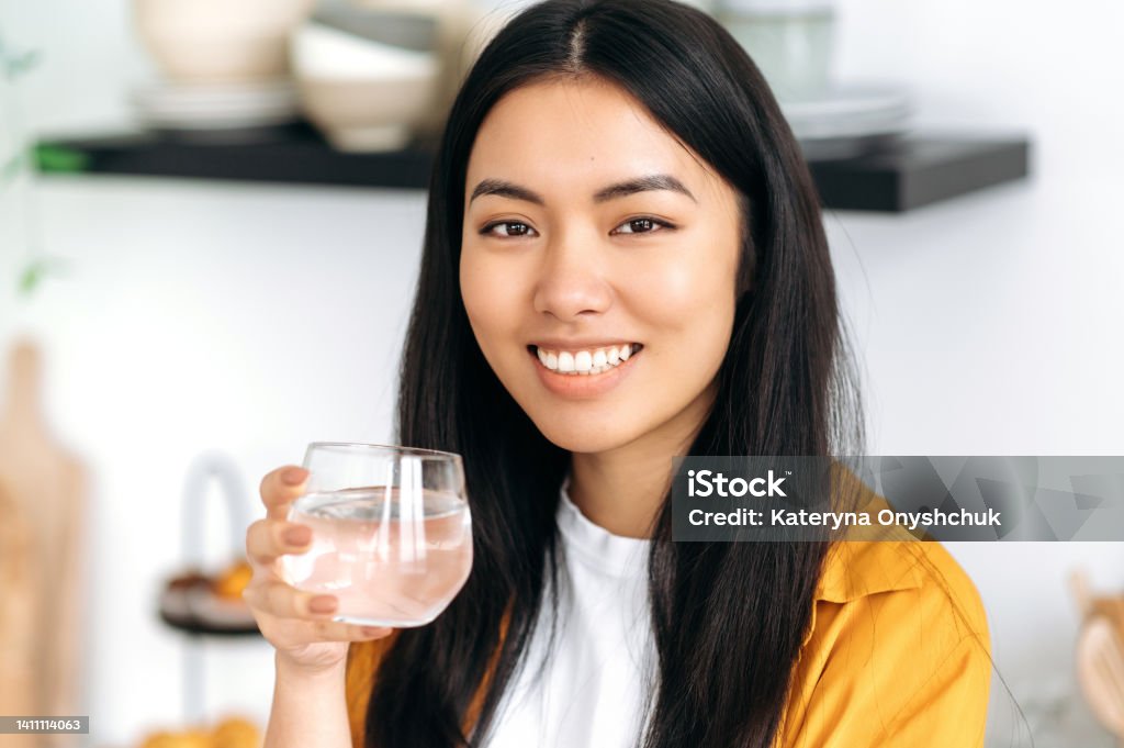 Pretty, healthy chinese brunette girl, dressed in casual stylish clothes, holds a glass of pure water, takes care of her health, drinks daily norm of water, looking at camera, smiles friendly 20-24 Years Stock Photo
