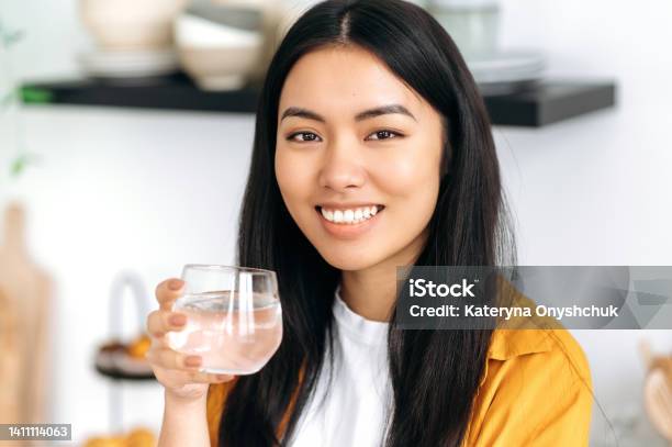 Pretty Healthy Chinese Brunette Girl Dressed In Casual Stylish Clothes Holds A Glass Of Pure Water Takes Care Of Her Health Drinks Daily Norm Of Water Looking At Camera Smiles Friendly Stock Photo - Download Image Now