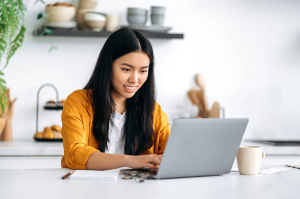 istock Positive friendly smiling Chinese girl, freelancer, programmer, manager, sitting at the table, working from home, using a laptop, working on a project with concentration, thinking over a strategy 1411114093