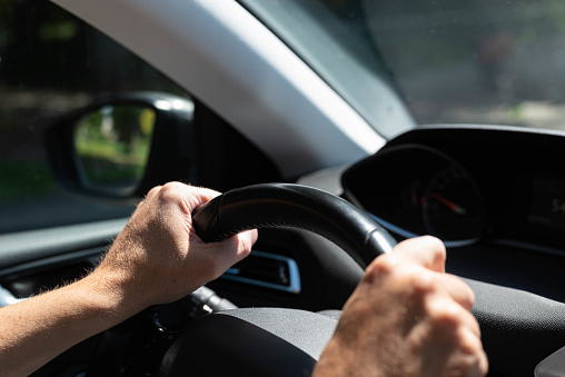 White men hands control a car steering wheel while driving on the road. There are no recognizable persons or trademarks in the shot.