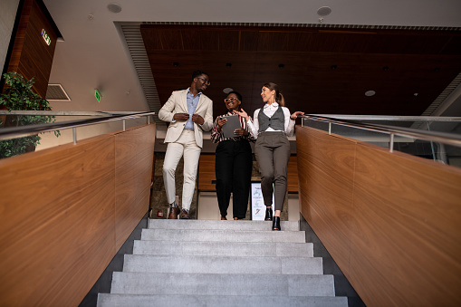 Three young business people walking down the staircase in a hotel. Three young interracial coworkers walking together on the staircase in a hotel. Group of young mixed race colleagues on a casual meeting. Interracial group of businesspeople.