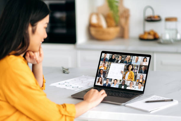Remote lesson. Positive smart Asian female student, sitting at the table, listening to an online lecture, on the screen of a laptop, a teacher telling information, and a group of multiracial people stock photo