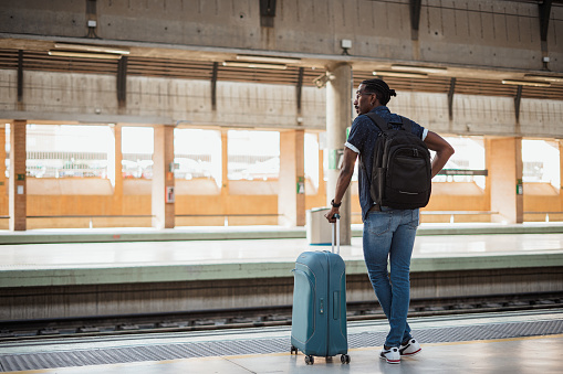 African male standing next to a platform while holding a blue suitcase. He is at a train station