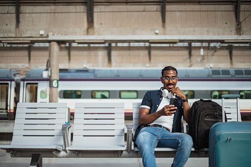 African male using his phone while is sitting on a bench at the train station.