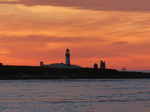 Viewed from the Pentland Firth, the Cantick Head Lighthouse on Switha Island is silhouetted by a bright orange sky at sunset.  Switha is an islet in Scotland’s Orkney archipelago.